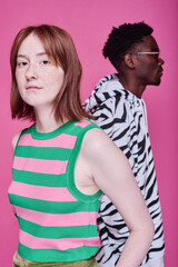 Portrait of red haired teenage girl looking at camera with African teenage boy standing behind her over pink background