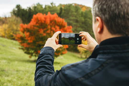 A man takes a photo of a beautiful landscape, a tree in an autumn park for his blog. Travel and outdoor activities.