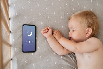 Upper view of baby napping in his bed next to phone playing lullaby in app for baby sleep with...