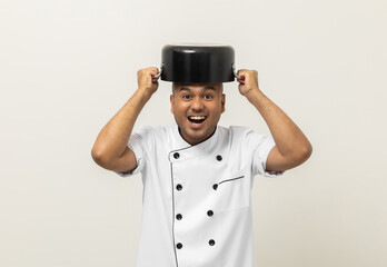 Funny excited face Young asian man chef in uniform put a stew pot over the head kitchen utensils on isolated. Cooking indian man Occupation chef People in kitchen restaurant and hotel.