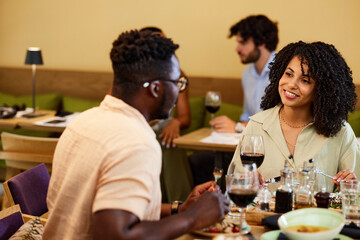A happy multiracial couple sits in a restaurant, chatting and having dinner together.