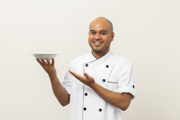 Smiling Young handsome asian man chef in uniform holding empty plate delicious dish menu good taste...
