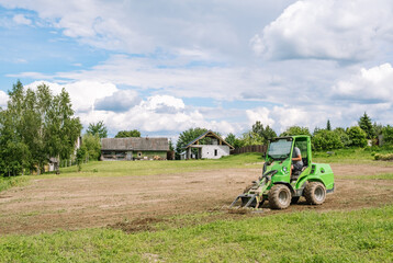 A green mini skid steer loader clear the construction site. Land work by the territory improvement. Small tractor with a ground leveler for moving soil, turf. Machine for agriculture work. Copy space