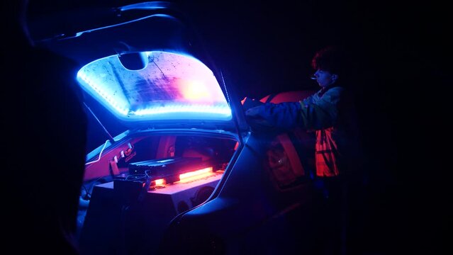 Young Man With Cigarette In Mouth Setting Up Car Stereo And Lighting In Rear Compartment. Illegal Rave Party In The Street At Night. wide