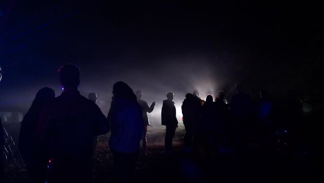Silhouette Of Party People Dancing In The Street At Night With A Car Arriving At The Scene. Street Gabbers In An Illegal Rave Party. wide establishing shot