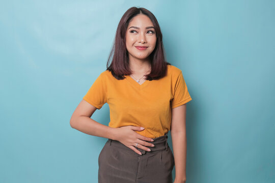 Pleased cheerful Asian woman keeps hand on belly feels full after delicious dinner dressed casually stands thoughtful against blue background.