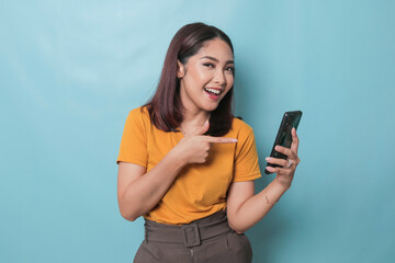 An excited young woman pointing her smartphone in her hand, isolated on blue background