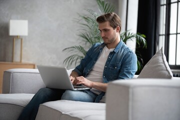 Young attractive smiling man is browsing at his laptop, sitting at home on the cozy sofa at home, wearing casual outfit.