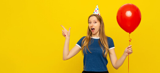 Young beautiful woman in a birthday cap is pointing at left banner with pleased tempting expression, showing advertisement, holds red balloon. Indoor studio shot on yellow background