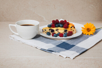 A piece of blueberry cheesecake on a white plate with fresh raspberries and blueberries and a cup of coffee on a stone table. Front view