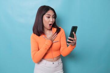 A surprised young woman pointing at copy space on her smartphone in her hand, isolated on blue background