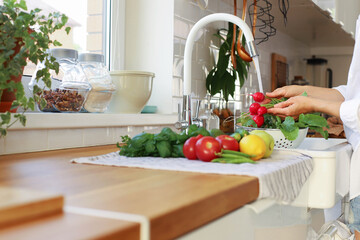 A young girl washing vegetables and fruits in the kitchen in the sink.