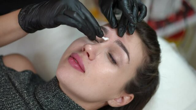 High angle view woman with closed eyes lying in beauty salon as unrecognizable beautician erasing contour leftovers from face. Close-up confident Caucasian beautiful client doing permanent makeup