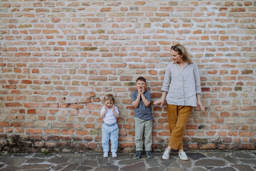 Young mother with her little children standing by brick wall and making funny faces in street...