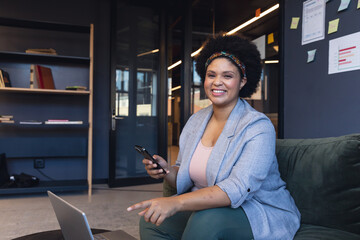 Portrait of smiling mid adult biracial businesswoman with smart phone sitting in office - Powered by Adobe