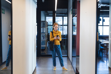 Mid adult caucasian businesswoman using mobile phone while standing in corridor of creative office