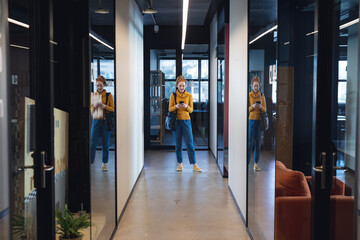 Mid adult caucasian businesswoman using smartphone while standing in corridor of creative office - Powered by Adobe