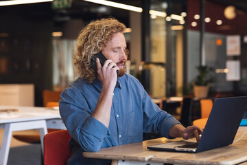Young caucasian businessman using laptop and using smart phone at desk in creative office