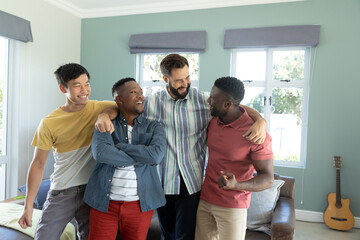 Smiling multiracial male friends with arms around talking while standing in living room at home