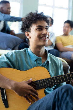 Smiling young man playing guitar while multiracial male friends relaxing in background at home