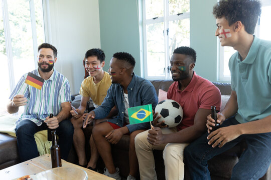 Happy multiracial male friends with flags, beer bottles, balls watching match while sitting on sofa