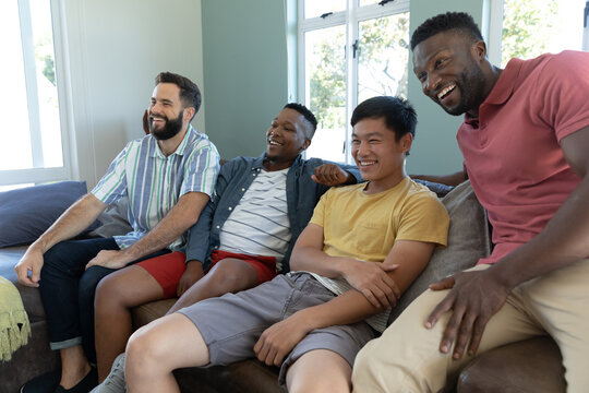 Happy multiracial male friends looking away and laughing while relaxing on sofa in living room