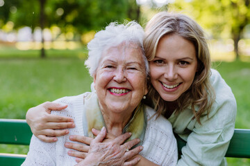 Portrait of adult granddaughter hugging her senior grandmother when sitting on bench in park.
