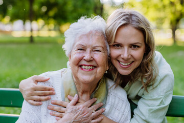 Portrait of adult granddaughter hugging her senior grandmother when sitting on bench in park. - Powered by Adobe