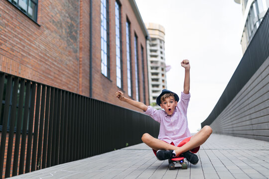 Caucasian Boy Sitting In Skateboard And Going Down The Hill, Looking At Camera.