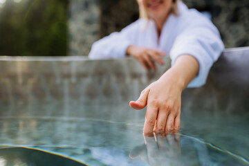 Unrecognizable woman in towel touching water, checking temperature, ready for home spa procedure in hot tub outdoors. Wellness, body care, hygiene concept.