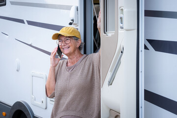 Smiling senior woman in travel vacation outside a camper van enjoying freedom and travel. Attractive elderly caucasian lady in van life standing at the door of motor home talking on mobile phone