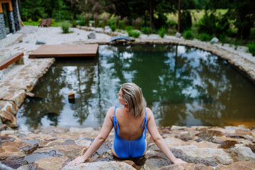 Rear view of young woman in swimsuit sitting by backyard natural pond during summer vacation in mountains.