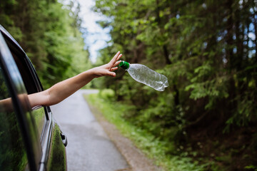 Woman's hand throwing away plastic bottle from car window on the road in green nature,...