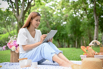 Fototapeta premium Happy young Asian female using tablet while picnicking in the green garden in summer day.