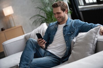 Attractive smiling young man wearing casual clothes sitting on a couch at the living room, using...