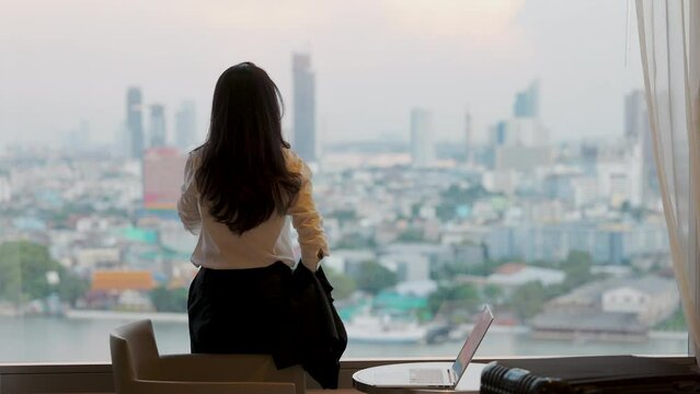 Relaxation holiday vacation of businesswoman take it easy happily resting standing nearly window looking at city scenery on the background with her luggage, laptop in hotel bedroom after check-in.