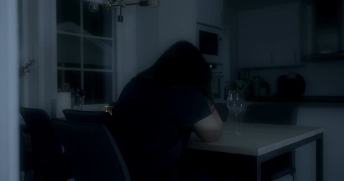 Back view of depressed drunk woman sitting at kitchen table at night. Slow pan right