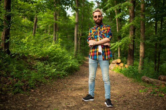 Full length image of a handsome young man with blonde hair, wear sunglasses, posing outdoors in the countryside, with crossed hands.