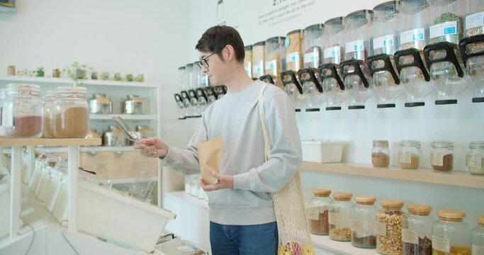 Man fill up reusable paper bag with bio organic product in zero waste shop. Asian male buying food in plastic free grocery store. Eco friendly business startup go green market. Reduce Reuse Recycle.
