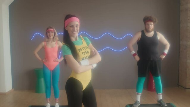 Portrait Of Dark-haired Caucasian Female Instructor Showing Her Physique, Young Man And Women Standing With Hands On Waist In 80s Aerobics Class Studio At Daytime, Looking On Camera