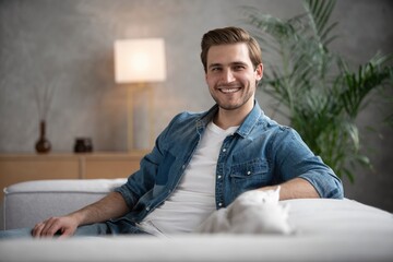 attractive caucasian man in denim shirt sitting on a sofa in the living room looking at the camera...