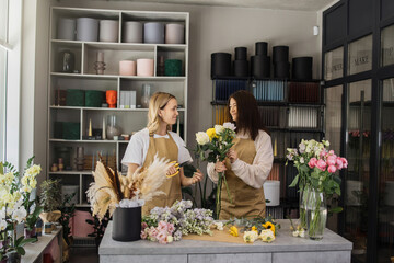Two smiling florists in uniform work in a flower shop behind counter with different varieties of...