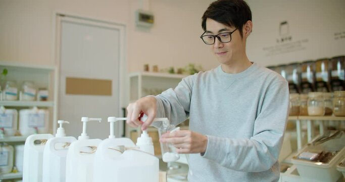 Young man bring his own containers buying eco bio organic goods in zero waste shop. Asian male refilling natural biodegradable household product from container dispenser in plastic free grocery store.