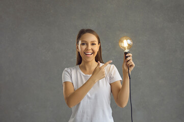 Cheerful smart and intellectual woman points to bright light bulb that she holds in her hand....