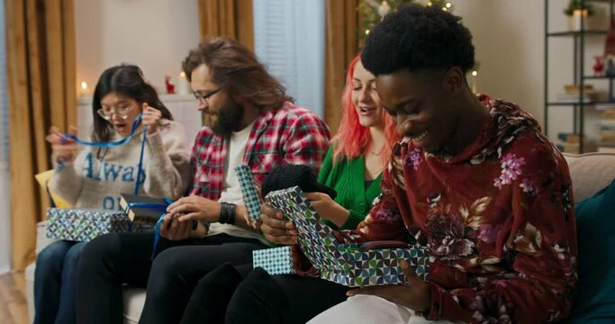 Unpacking gifts is the most magical moment at Christmas party. Smiling friends sit on the sofa in the living room and rejoice in new gifts. Boys and girls smile and discuss the gift.
