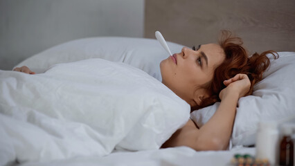 redhead woman measuring temperature with electronic thermometer and lying on bed.