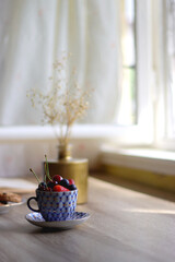 Plate of chocolate chip cookies, cup filled with strawberries, blueberries and cherries, open book and vase with gypsophila flowers on the table. Selective focus.