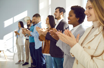 Joyful and satisfied audiences applaud speaker during business conference or seminar. Side view of smiling multiracial people standing in row and applauding looking in same direction. Selective focus.