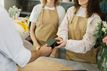 Close up of caucasian male hands using electronic credit card on his smartphone paying for purchase using payment terminal in flower shop. Client buying bouquet making payment in store