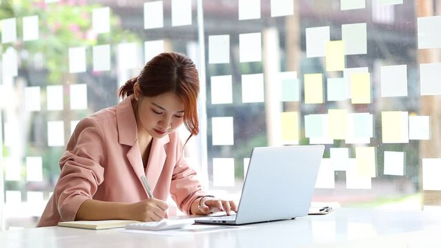 Attractive Asian woman working on laptop and writing work notes.